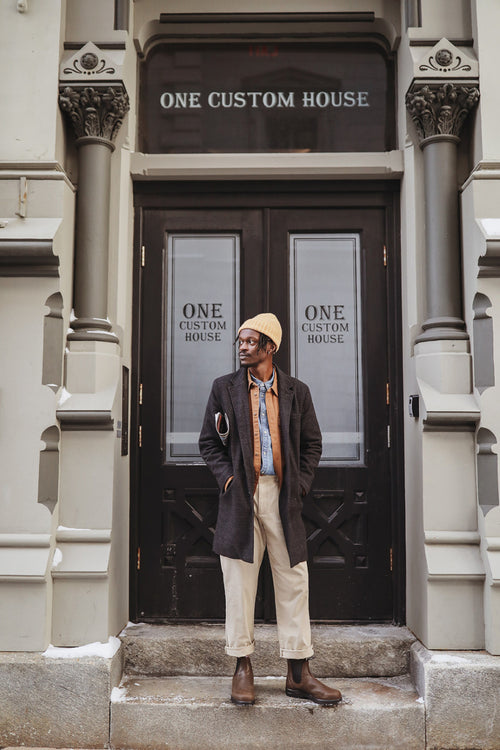 A person wearing a yellow beanie, glasses, dark coat, blue shirt, tan pants, and brown boots stands on stone steps in front of double doors labeled “One Custom House.â€.