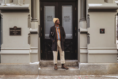 A person in a dark coat, beige pants, brown boots, and a yellow beanie stands in front of a double door labeled One Custom House on a city street with light snow on the ground.