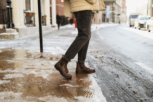 Person wearing dark jeans and brown boots steps onto a snowy, icy sidewalk from a road in an urban area. The scene suggests cold winter weather.