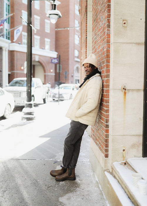 A person in a light-colored coat, beanie, and boots leans against a brick wall on a sunny, snowy city street, smiling. Buildings, cars, and street signs are visible in the background.