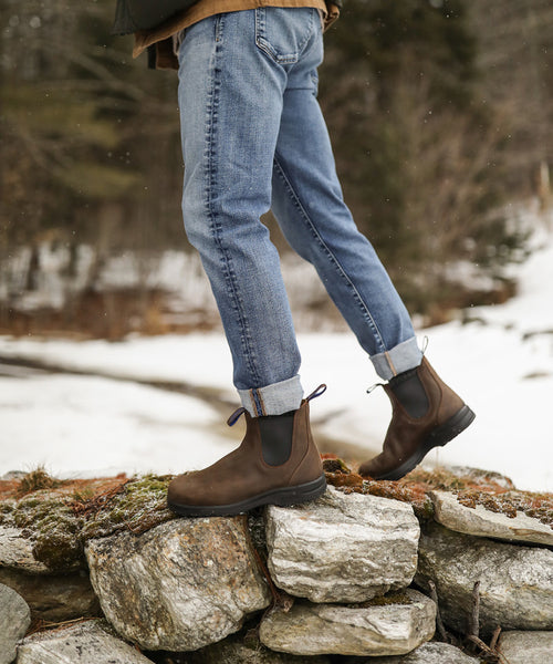 A person wearing cuffed blue jeans and brown boots stands on a stone wall outdoors with snow and trees in the background.