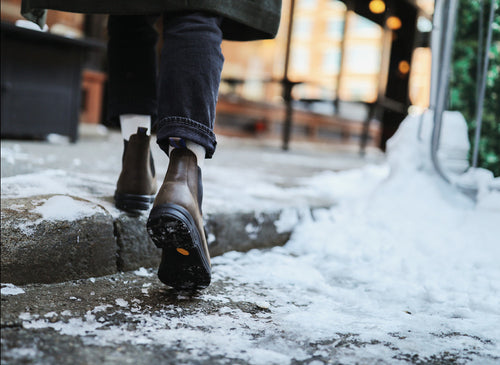 Person wearing brown boots and black pants walking on a snowy, icy sidewalk, viewed from behind at ground level. Snow and patches of ice are visible on the ground.