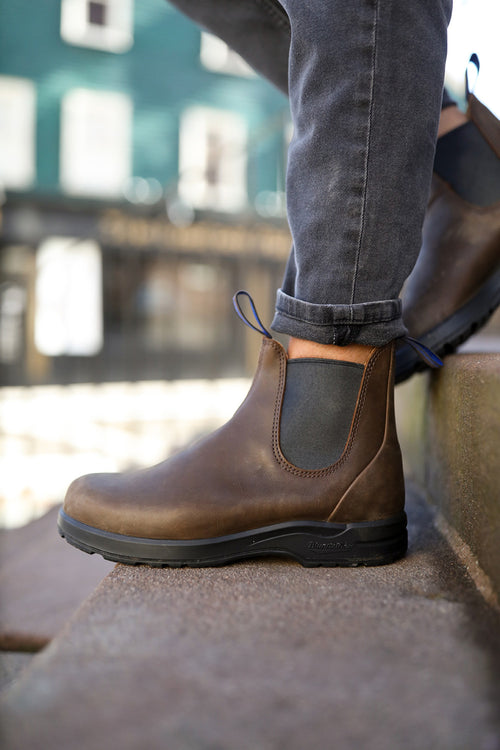 A person wearing dark jeans and brown leather Chelsea boots stands on outdoor stone steps, with one foot on a higher step. The background is blurred, showing buildings.