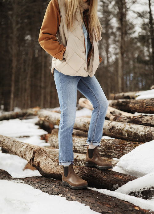 A person wearing a tan jacket, blue jeans, and brown boots stands on snow-covered logs in a forest with bare trees. Their face is not visible.