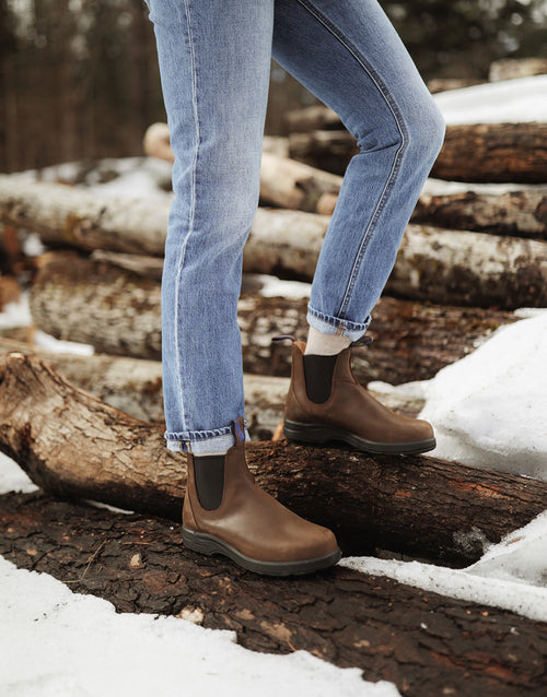 A person wearing brown Chelsea boots and rolled-up blue jeans steps on snow-covered logs in an outdoor, wooded area.
