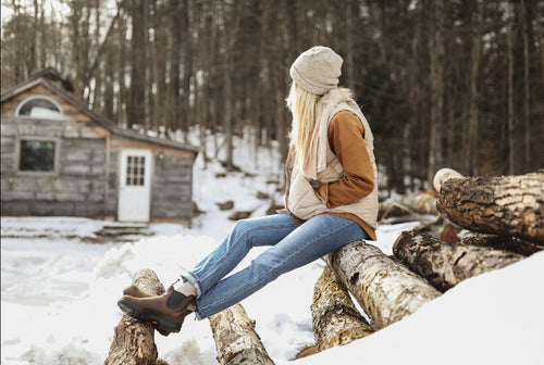 A person in a winter hat, coat, and boots sits on snowy logs, looking toward a rustic wooden cabin surrounded by bare trees.