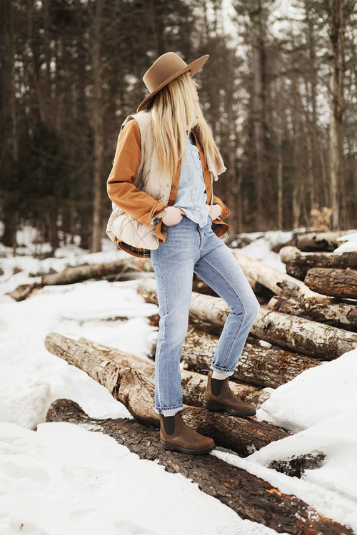 A woman wearing a brown hat, puffer vest, denim shirt, jeans, and brown boots stands on snow-covered logs in a winter forest, looking to the side.