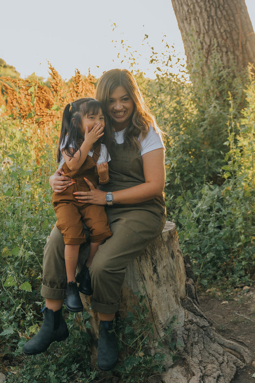 A woman and a young girl, both wearing brown overalls and white shirts, sit together on a tree stump outdoors, smiling and laughing in a lush, green setting.
