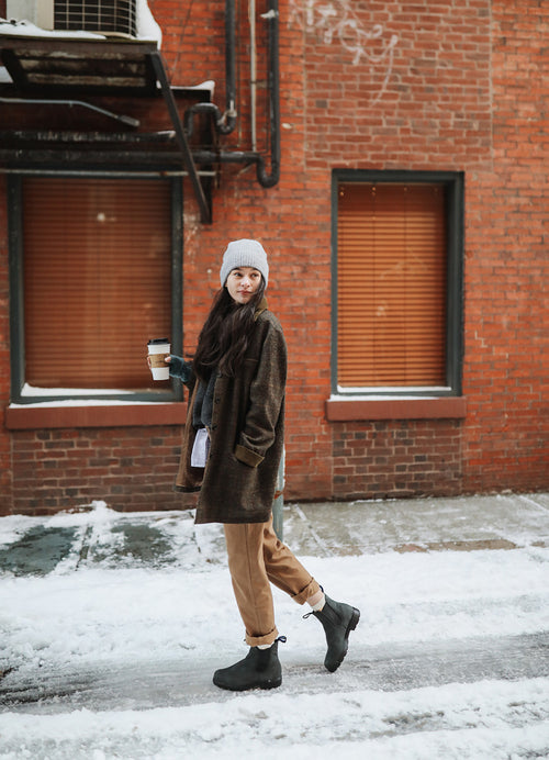 A woman wearing a gray beanie, brown coat, tan pants, and black boots walks on a snowy sidewalk, smiling and holding a coffee cup, in front of a red brick building with closed blinds.