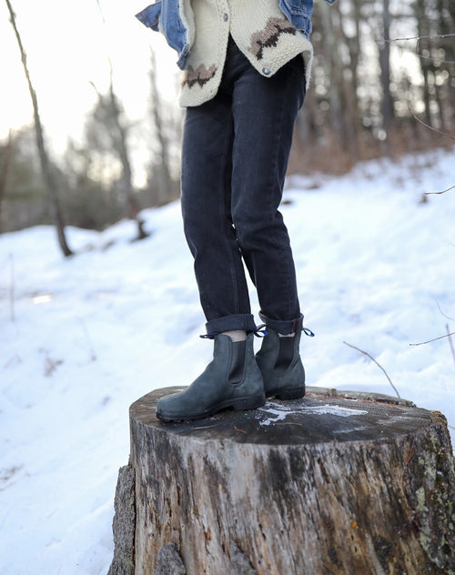 Person standing on a tree stump in a snowy forest, wearing black pants, dark boots, and a cream sweater with a denim jacket. Only the lower half of their body is visible.