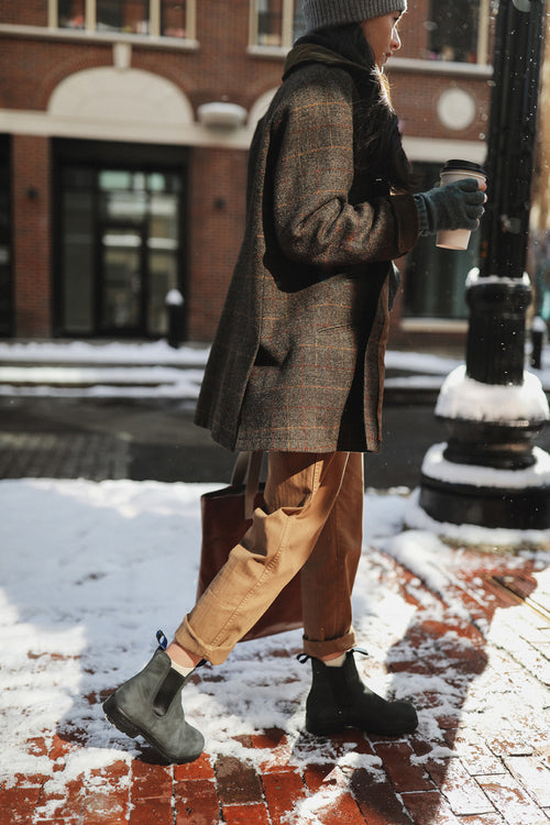 A person wearing a plaid coat, tan pants, black boots, and gloves walks on a snowy brick sidewalk while holding a coffee cup and a tote bag. A building and snow are visible in the background.