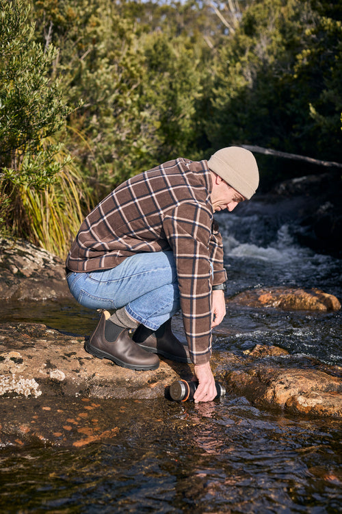A person in a beanie, plaid shirt, jeans, and boots crouches on a rock by a stream, filling a bottle with water. Green foliage surrounds the flowing water.