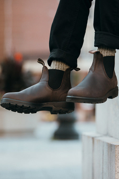 A person wearing dark brown Chelsea boots, gray wool socks, and black pants stands on a stone step outdoors, with one foot slightly raised. The background is softly blurred.