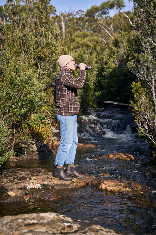 A person wearing a beanie, plaid shirt, jeans, and boots stands on rocks by a flowing creek, looking through binoculars amid dense, green bushland.