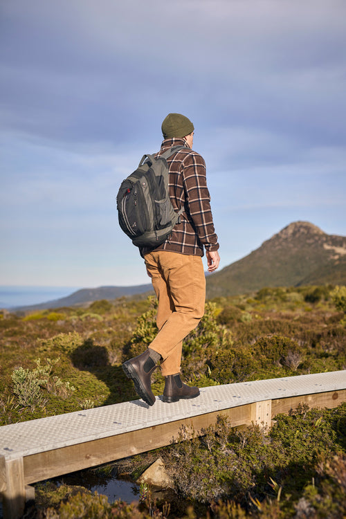 A person wearing a beanie, plaid shirt, tan pants, and boots walks on a raised boardwalk through a grassy landscape with mountains in the background, carrying a black backpack.