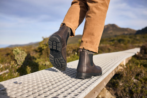 A person wearing brown leather boots and rolled-up tan pants walks along a raised metal boardwalk outdoors, surrounded by greenery and distant mountains under a blue sky.