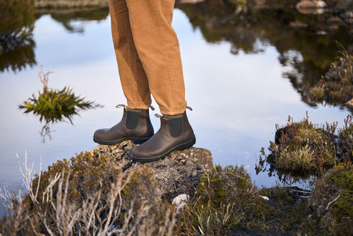 A person wearing brown pants and dark brown boots stands on a rock surrounded by water and vegetation in an outdoor setting.