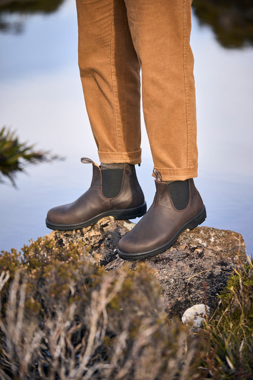 A person wearing brown Chelsea boots and tan pants stands on a rocky surface near water, with green foliage and moss visible in the foreground.
