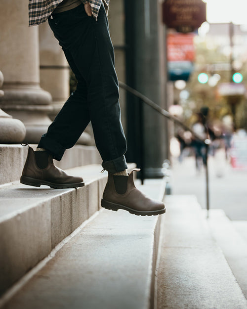 A person wearing dark pants and brown Chelsea boots walks up concrete steps outside, with buildings and blurred pedestrians in the background.
