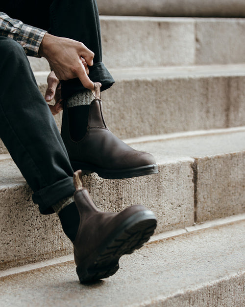A person sitting on concrete steps puts on brown leather Chelsea boots, wearing dark pants with cuffs and grey socks. Only the lower body and hands are visible.