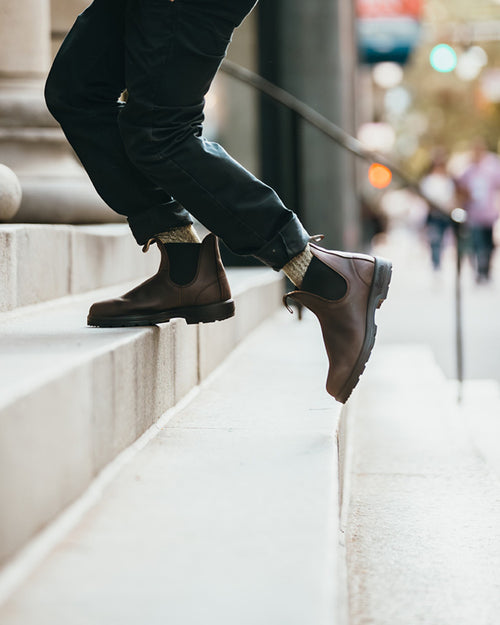 Person wearing dark pants and brown Chelsea boots walking up outdoor stone steps, with one foot raised and blurred city lights in the background.