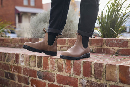 A person wearing brown Chelsea boots and black pants stands on a red brick wall, with plants and a building visible in the background.