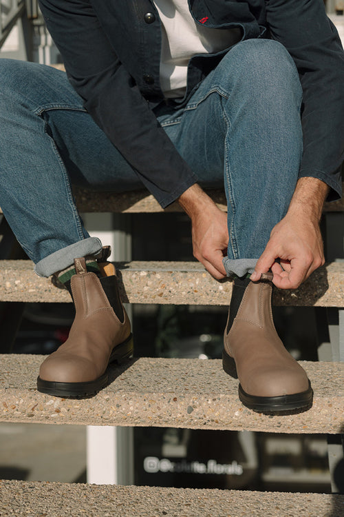 A person in rolled-up blue jeans and a dark jacket sits on outdoor steps, putting on light brown Chelsea boots.