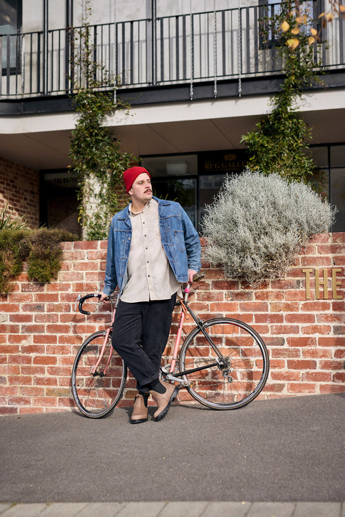 A man wearing a red beanie, denim jacket, beige shirt, and black pants leans against a pink bicycle in front of a brick wall with greenery. An apartment building with balconies is in the background.