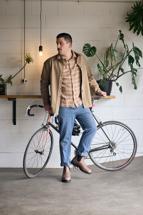 A man wearing a beige jacket, plaid shirt, and jeans leans against a bicycle indoors, with plants and a hanging lightbulb behind him on a white brick wall.