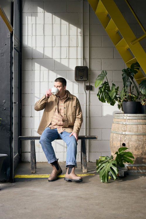 A man in casual clothes sits on a bench indoors, drinking from a cup. He is surrounded by potted plants and a wooden barrel, with sunlight streaming through a window and a yellow staircase in the background.