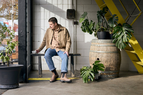 A man sits on a bench near a large window, holding a cup. He wears jeans, boots, a plaid shirt, and a tan jacket. Nearby are potted plants, a wooden barrel, and yellow stairs against a white brick wall.