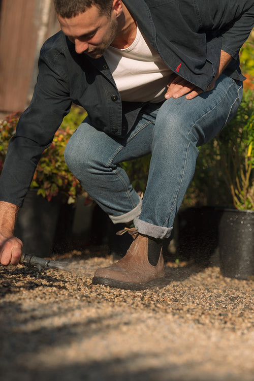 A man in jeans, boots, and a jacket crouches outdoors, using a small garden tool to work on gravel near rows of potted plants in sunlight.