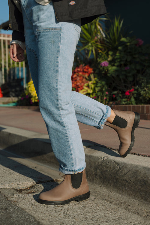 A person in light blue jeans and tan Chelsea boots stands on a curb, with one leg bent and foot lifted behind. Colorful plants and flowers are visible in the background.