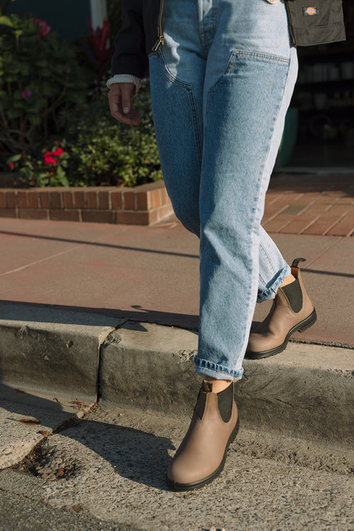 A person wearing light blue jeans and taupe Chelsea boots steps onto a curb on a sunny day, with greenery and flowers visible in the background.
