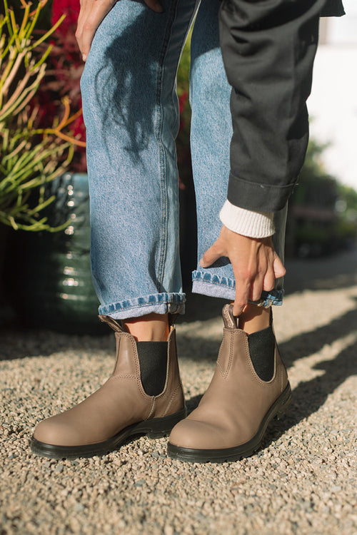 A person wearing cuffed blue jeans and taupe Chelsea boots stands on gravel, adjusting one boot. Green plants are visible in the background.