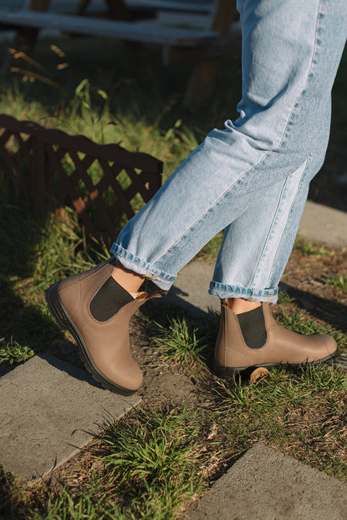 A person wearing light blue jeans and tan ankle boots with black elastic sides is walking outdoors on stone pavers with grass around them.
