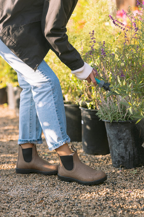 A person wearing brown boots, light blue jeans, and a black jacket bends down to cut or trim potted plants with scissors in an outdoor garden area.