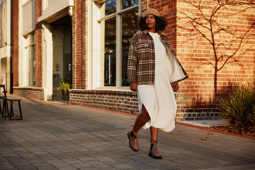 A woman in a plaid jacket, white dress, and tan beret walks on a paved sidewalk beside a brick building, with soft sunlight creating long shadows and highlighting her casual, stylish look.