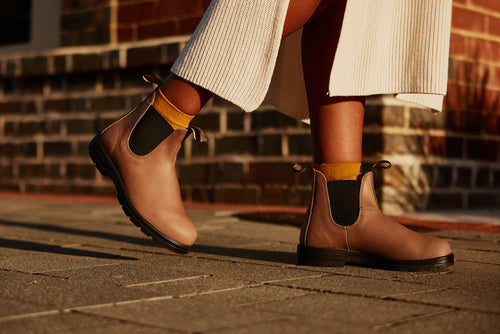 A person wearing tan Chelsea boots with yellow socks and a cream-colored ribbed skirt stands on a sunlit stone pavement in front of a brick wall.