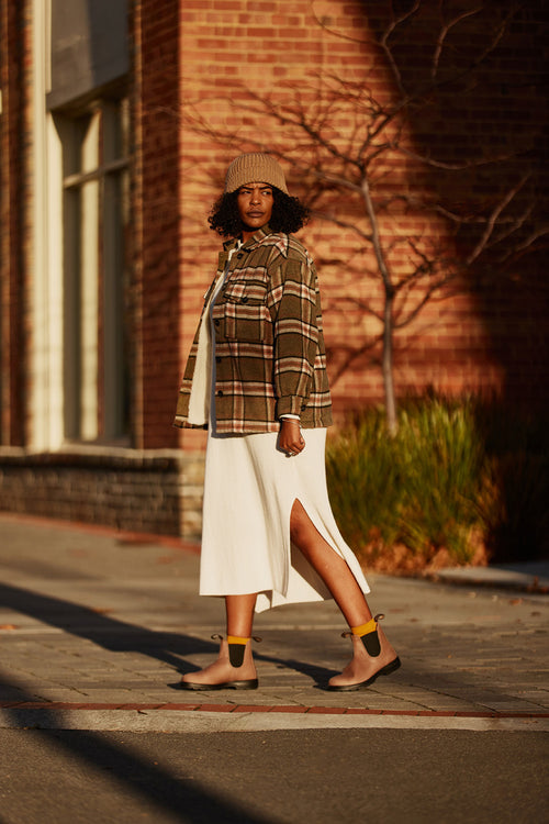 A woman in a tan beanie, plaid jacket, and white dress with a side slit walks on a sunlit sidewalk near a brick building, with bare branches and greenery in the background.