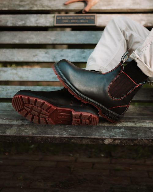 A person wearing light pants and black Chelsea boots with red stitching rests their feet on a wooden bench, showing the boots’ tread and relaxed posture.
