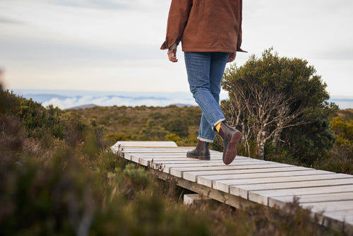 A person wearing jeans, brown boots, and a brown jacket walks away on a wooden boardwalk through a natural landscape with bushes and distant hills under a cloudy sky.