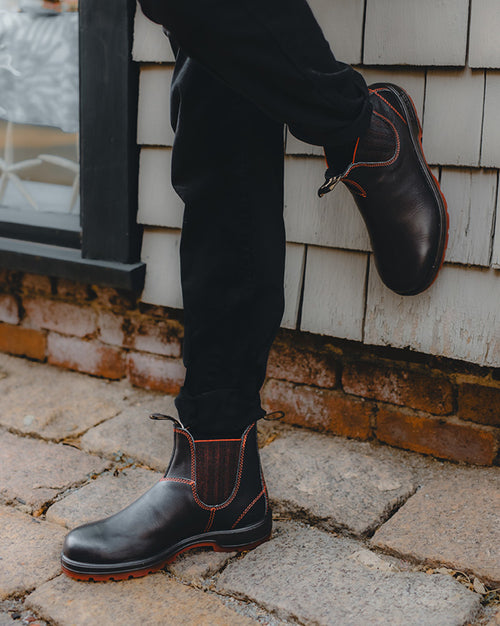 A person wearing black pants and dark brown leather Chelsea boots with red stitching stands on a stone walkway, one foot propped against a shingled wall and brick ledge.