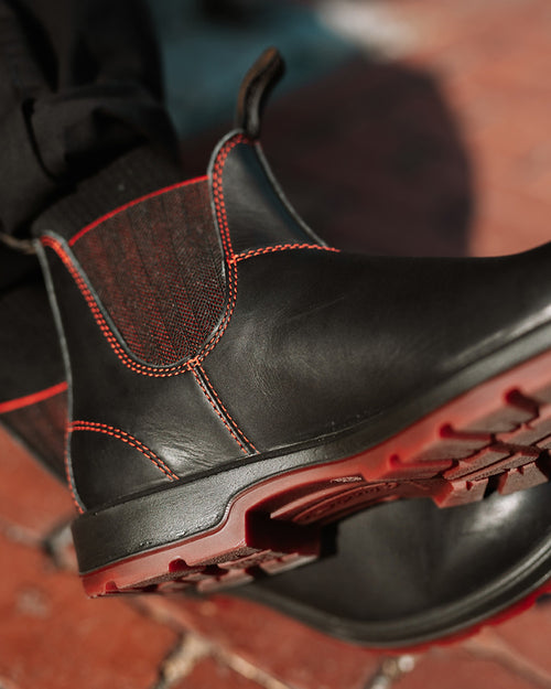Close-up of a black boot with red stitching and a red sole, worn by a person in black pants, standing on a reddish-brown brick surface.