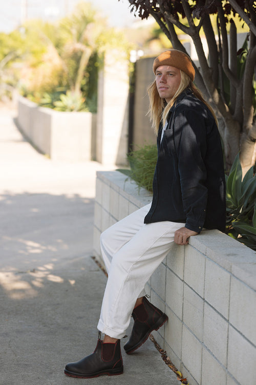 A person with long blond hair, wearing a mustard beanie, black jacket, white pants, and brown boots, sits on a low concrete wall outdoors near green plants and trees.