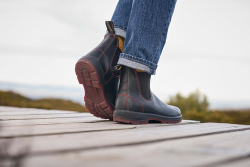 A person wearing dark brown Chelsea boots with red stitching and blue jeans stands on a wooden platform outdoors, with a blurred natural landscape in the background.