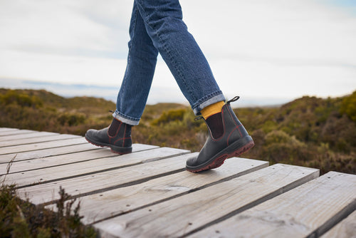 A person wearing jeans, yellow socks, and black boots walks on a wooden boardwalk outdoors, surrounded by bushes and natural landscape.