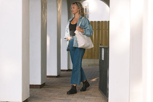 A woman wearing a denim jacket and jeans, black top, and black boots carries a large white tote bag while walking through a covered outdoor walkway with white columns.