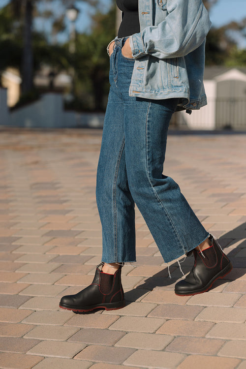 Person walking outdoors on a paved surface, wearing cropped blue jeans, a light denim jacket, and black ankle boots with red accents. The upper body is partially out of frame.