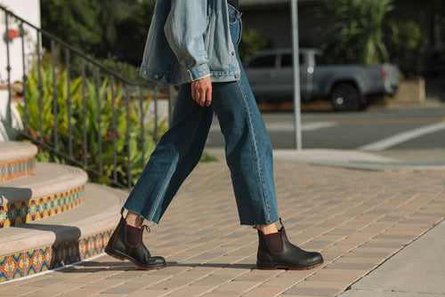 A person wearing a denim jacket, blue jeans, and black ankle boots walks on a paved sidewalk near decorative tiled steps and greenery, with a parked gray truck in the background.